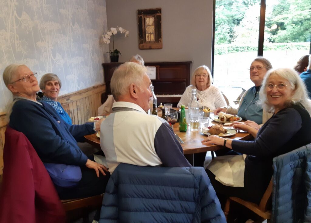 A group of seven sitting round a table at Daisy Clough cafe. A beautiful orchid is in the background and there is sun outside.
