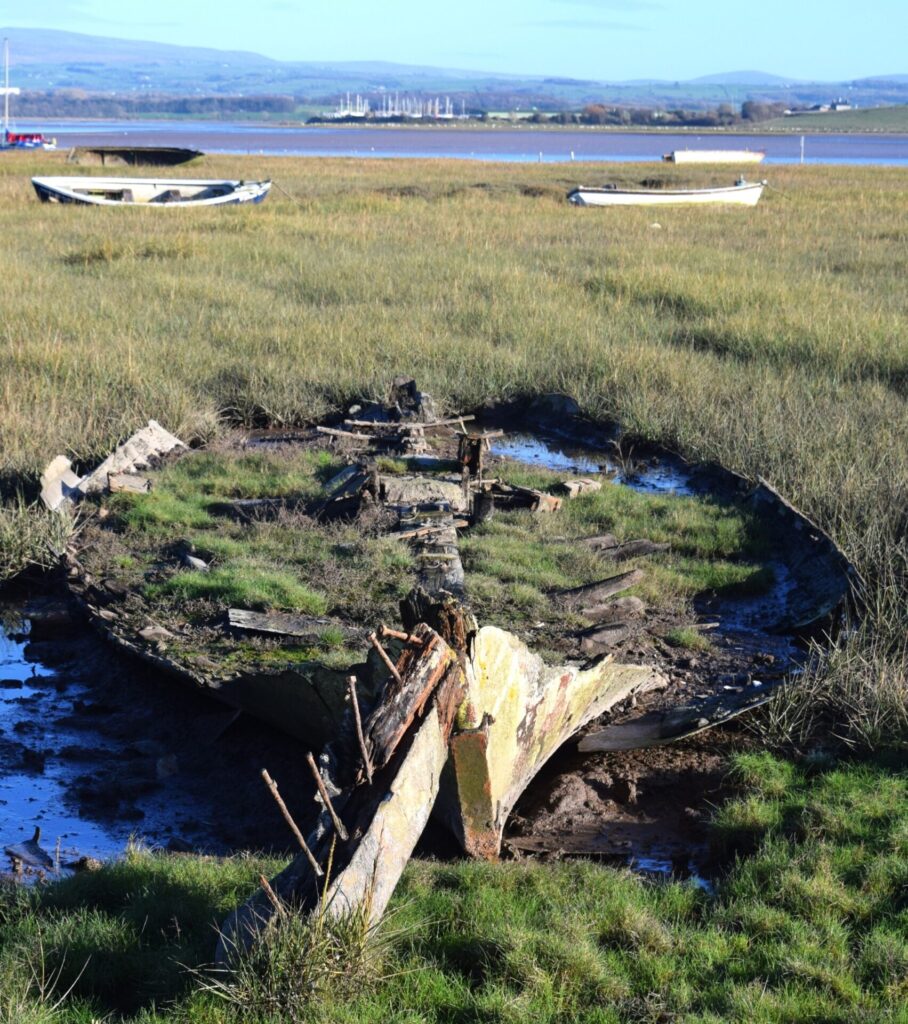 Sunderland Point. Old boat almost buried in the mud. Other boats in distance. Hills in the background.