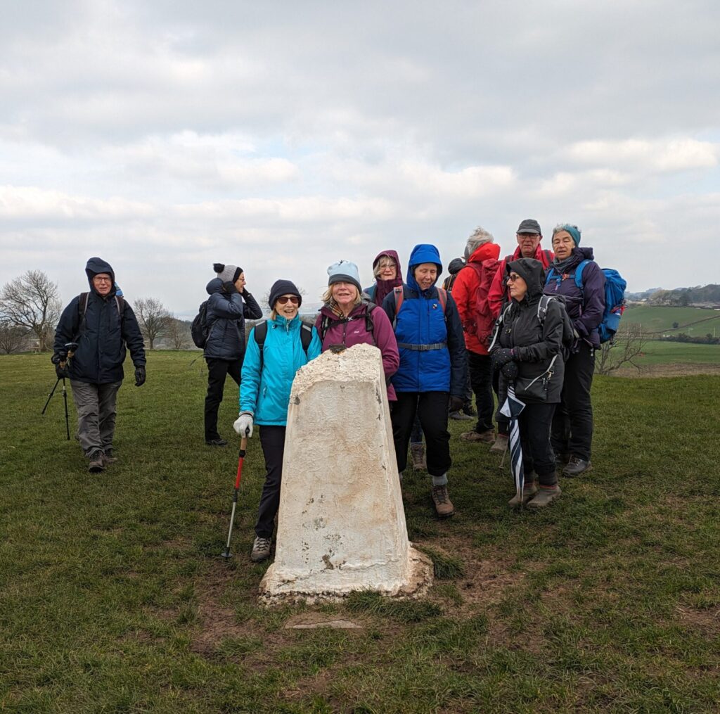 At a trig point  on a cold, cloudy day. The trig point is in the foreground and the top has broken off leaving a jagged edge.