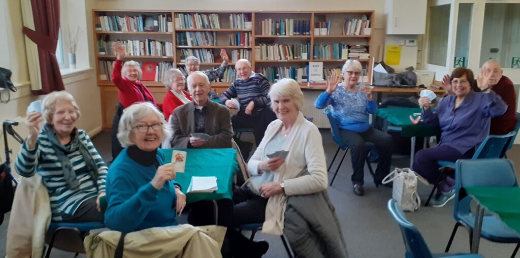 People sitting four to a table, smiling and waving at the camera.