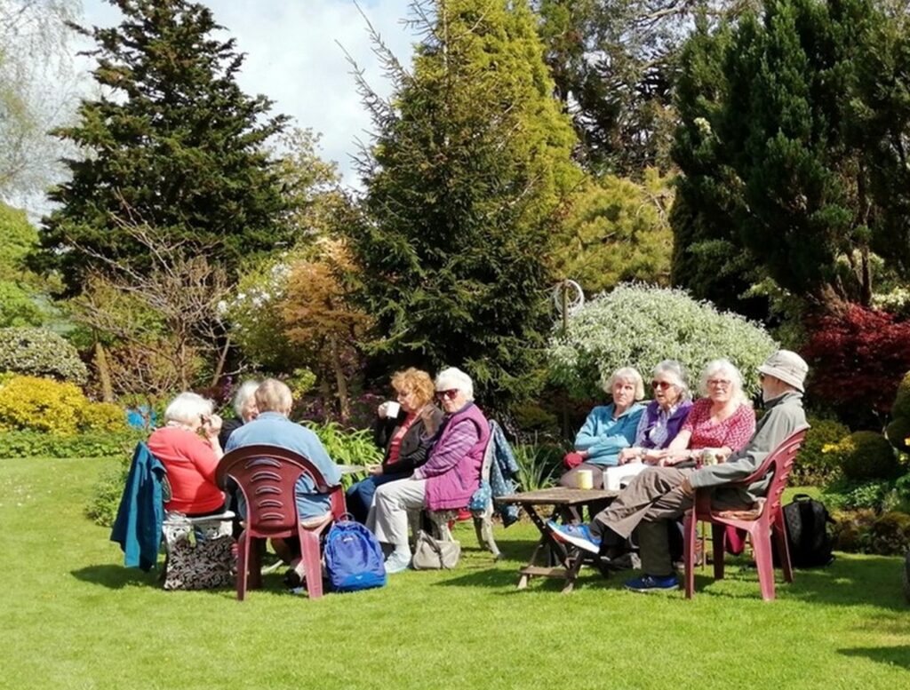 Members of the group visiting a garden which is part of the Open Gardens scheme. Sitting on chairs on the lawn, mixed shrubbery behind them making an array of different colours.