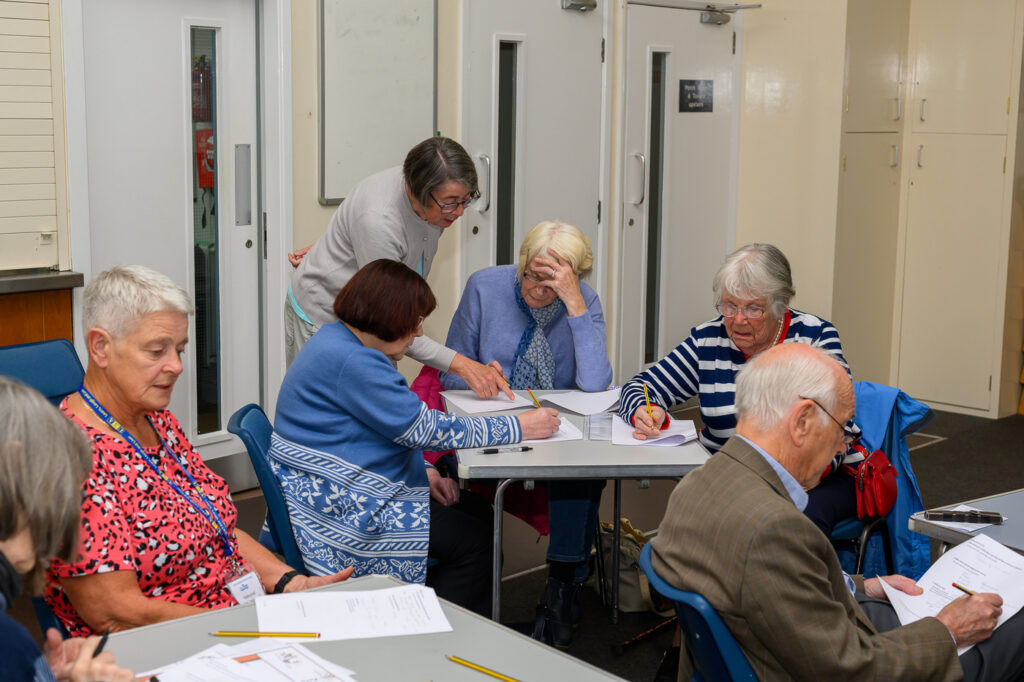 Being taught how to write a haiku at Japanese Day. Several people at tables with the Creative Writing Group Leader helping. Some head scratching.