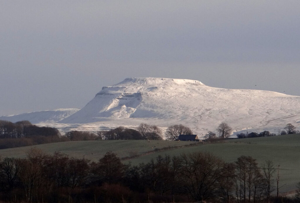 Ingleborough covered in snow though the fields in the foreground of the photo are green.