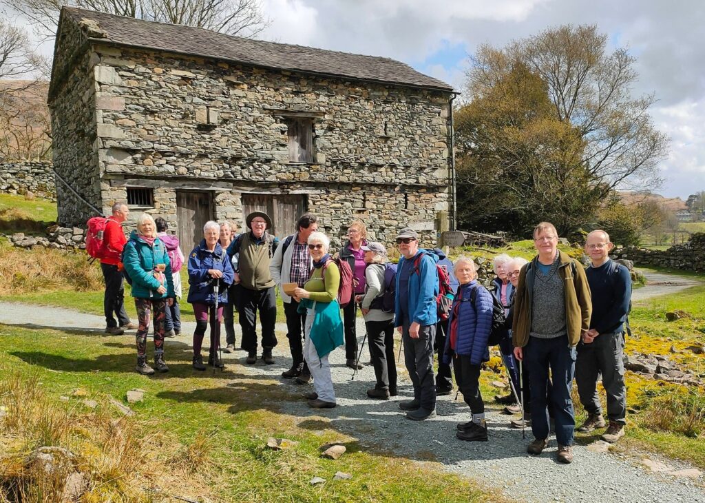A large group of walkers in the sun by an old barn.