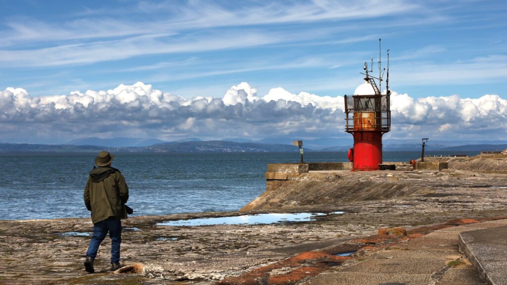 Beautiful blue sky, fluffy clouds right across the photo, fells in the distance. Red lighthouse in the foreground and a photographer.