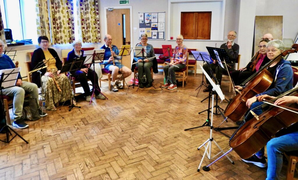 Members sitting in a circle with a wide range of instruments from cellos to recorders.